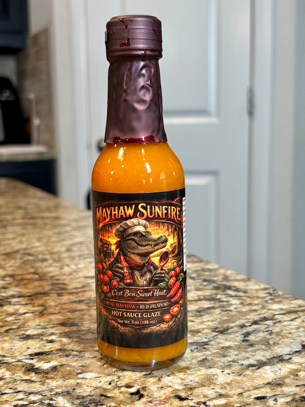 A bottle of Mayhaw Sunfire Red Hot Sweet Heat hot sauce glaze standing on a granite countertop with a kitchen background.