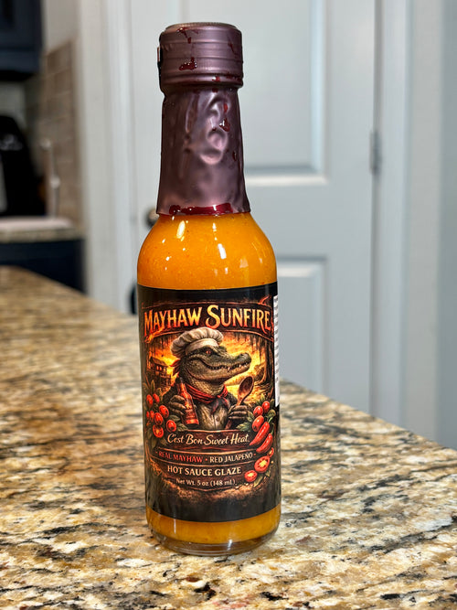 A bottle of Mayhaw Sunfire Red Hot Sweet Heat hot sauce glaze standing on a granite countertop with a kitchen background.