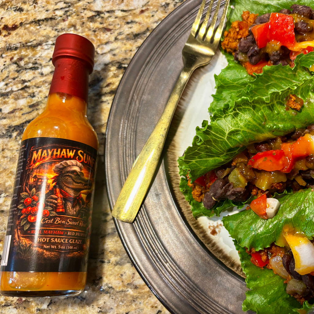 A bottle of Mayhaw Sunfire hot sauce placed beside a plate of food on a granite countertop. The plate holds lettuce wraps filled with beans, onions, and diced red peppers, with a gold fork resting on the plate.