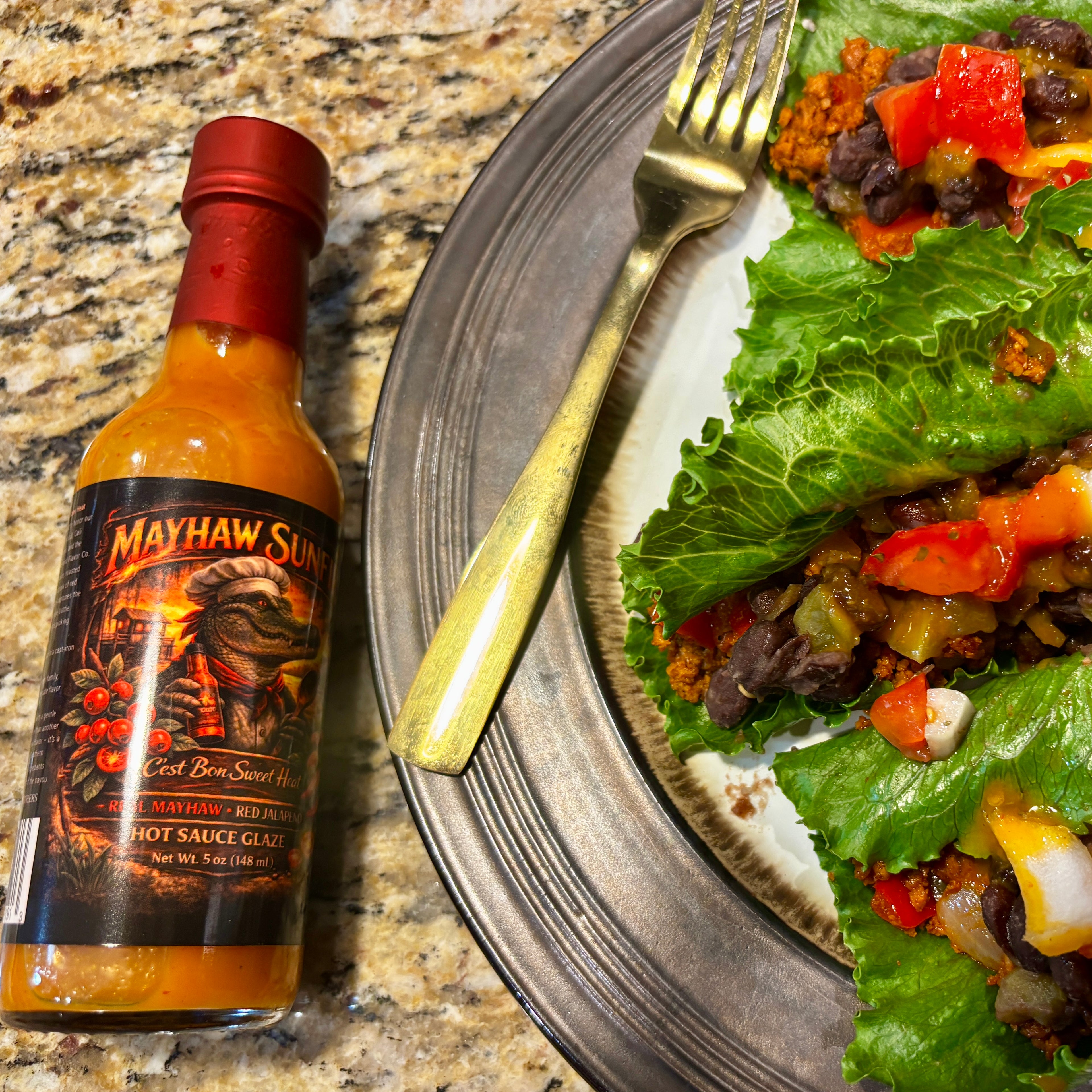 A bottle of Mayhaw Sunfire hot sauce placed beside a plate of food on a granite countertop. The plate holds lettuce wraps filled with beans, onions, and diced red peppers, with a gold fork resting on the plate.