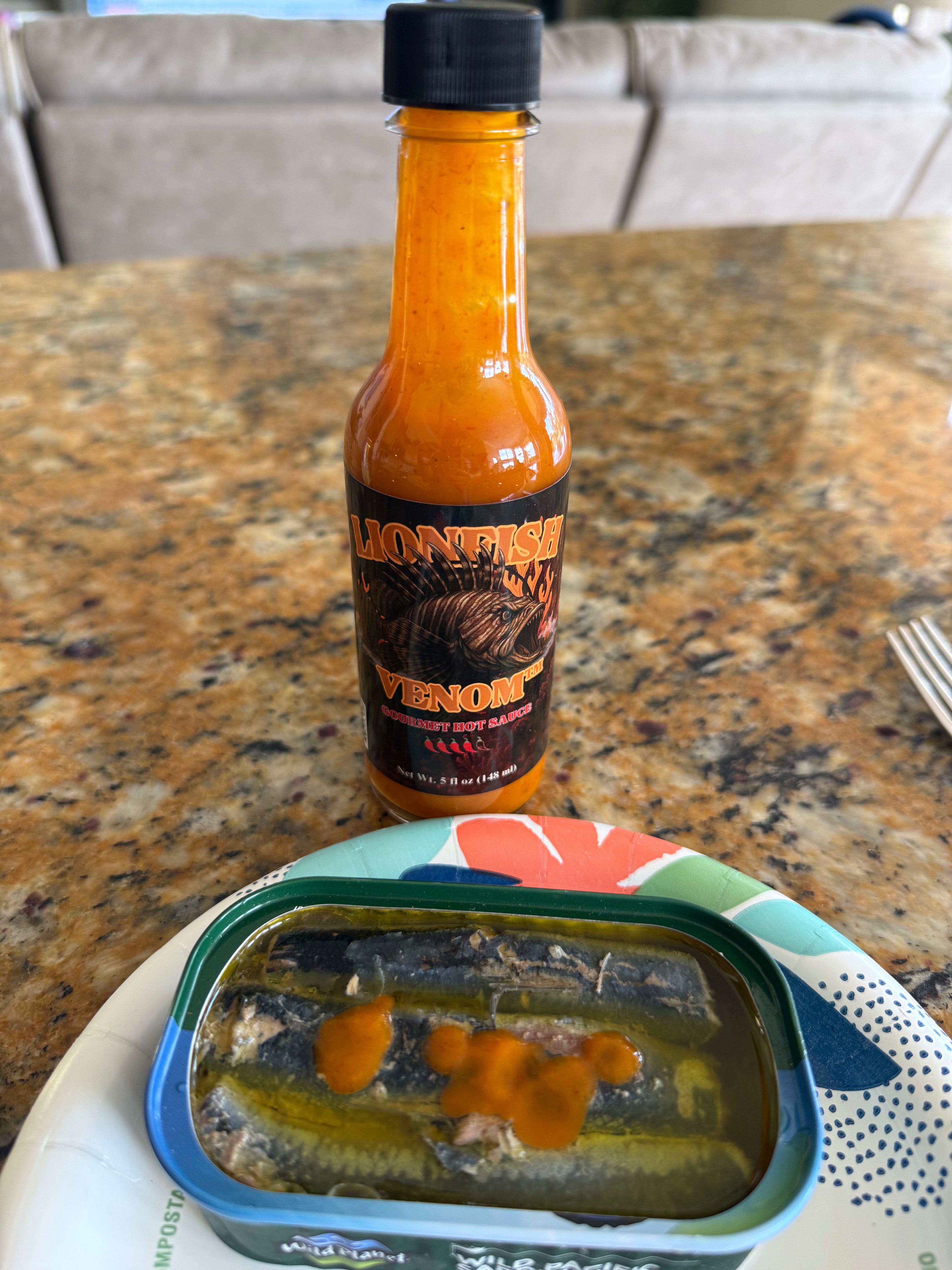 A bottle of Lionfish Venom hot sauce standing on a granite countertop next to an open can of mackerel fillets with hot sauce drizzled on top, with a patterned plate underneath.