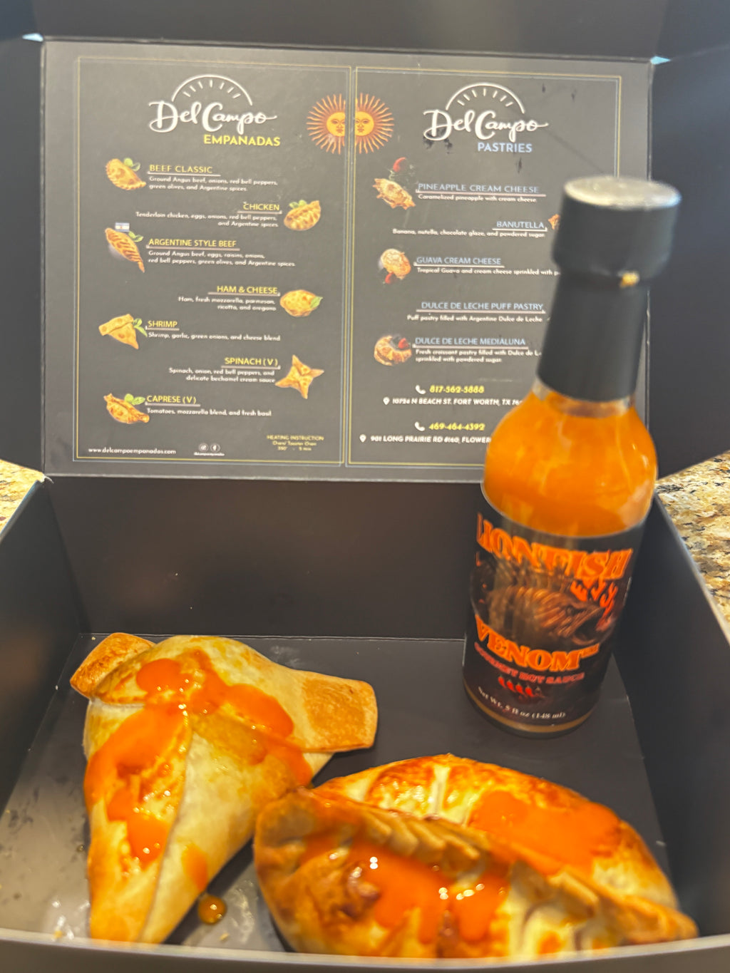 A black food delivery box containing two baked empanadas topped with orange sauce and a bottle of Del Corpo Habanero Pepper Sauce, with the open menu insert visible in the background.