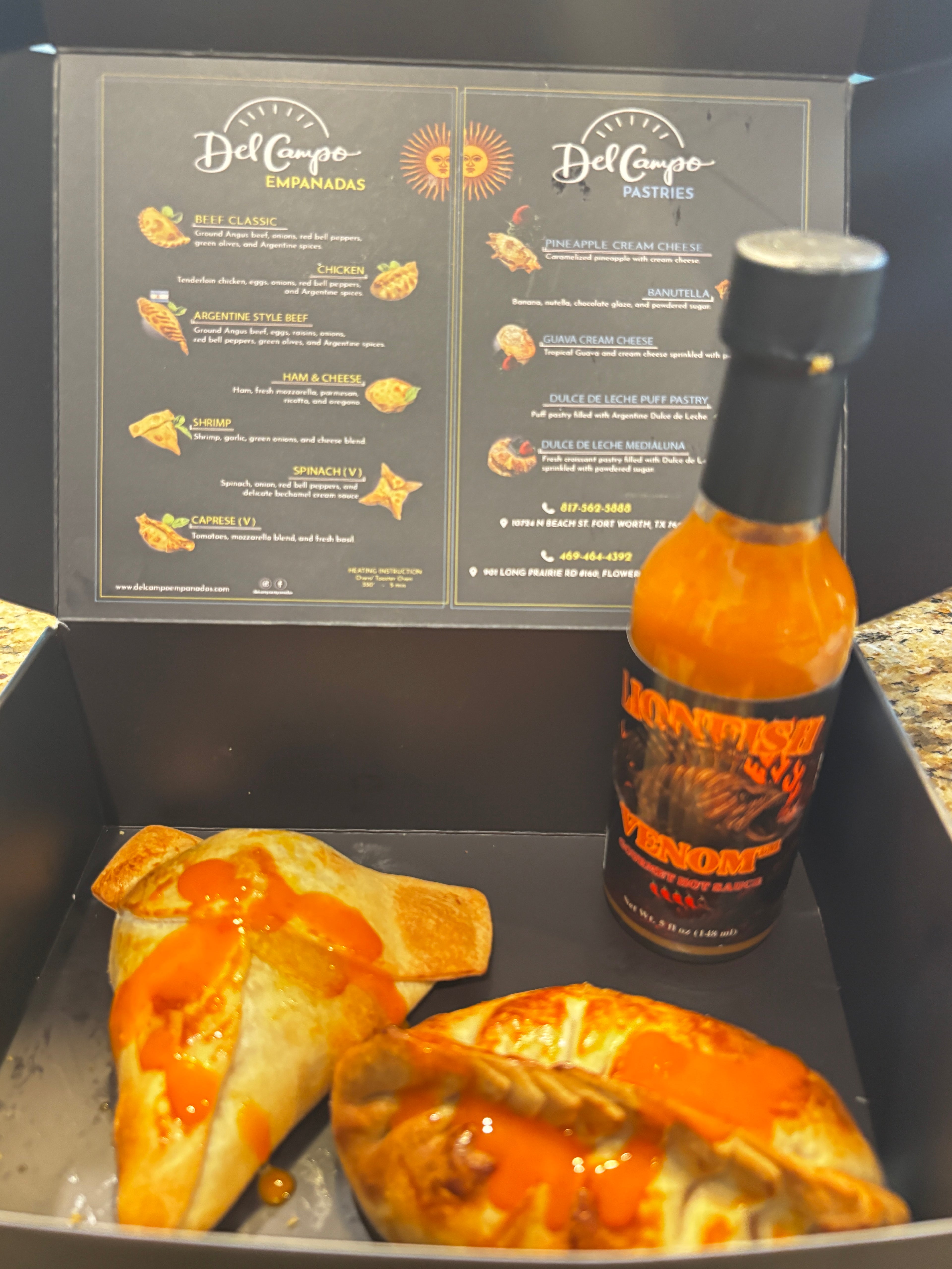 A black food delivery box containing two baked empanadas topped with orange sauce and a bottle of Del Corpo Habanero Pepper Sauce, with the open menu insert visible in the background.