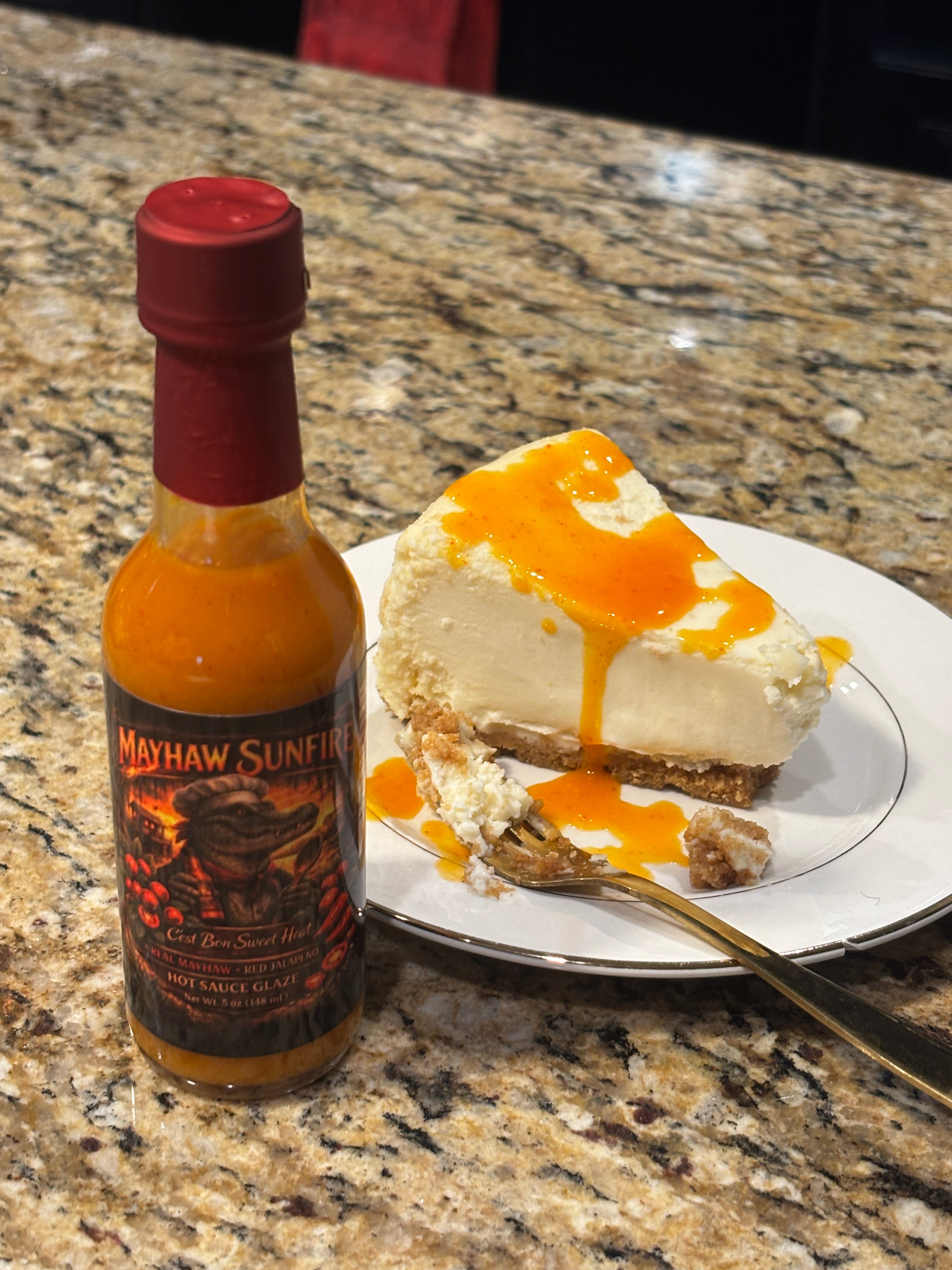  Mayhaw Sunfire hot sauce bottle stands beside a slice of vegan cheesecake drizzled with mayhaw sunfire hotsauce on a white plate, set on a granite counter 