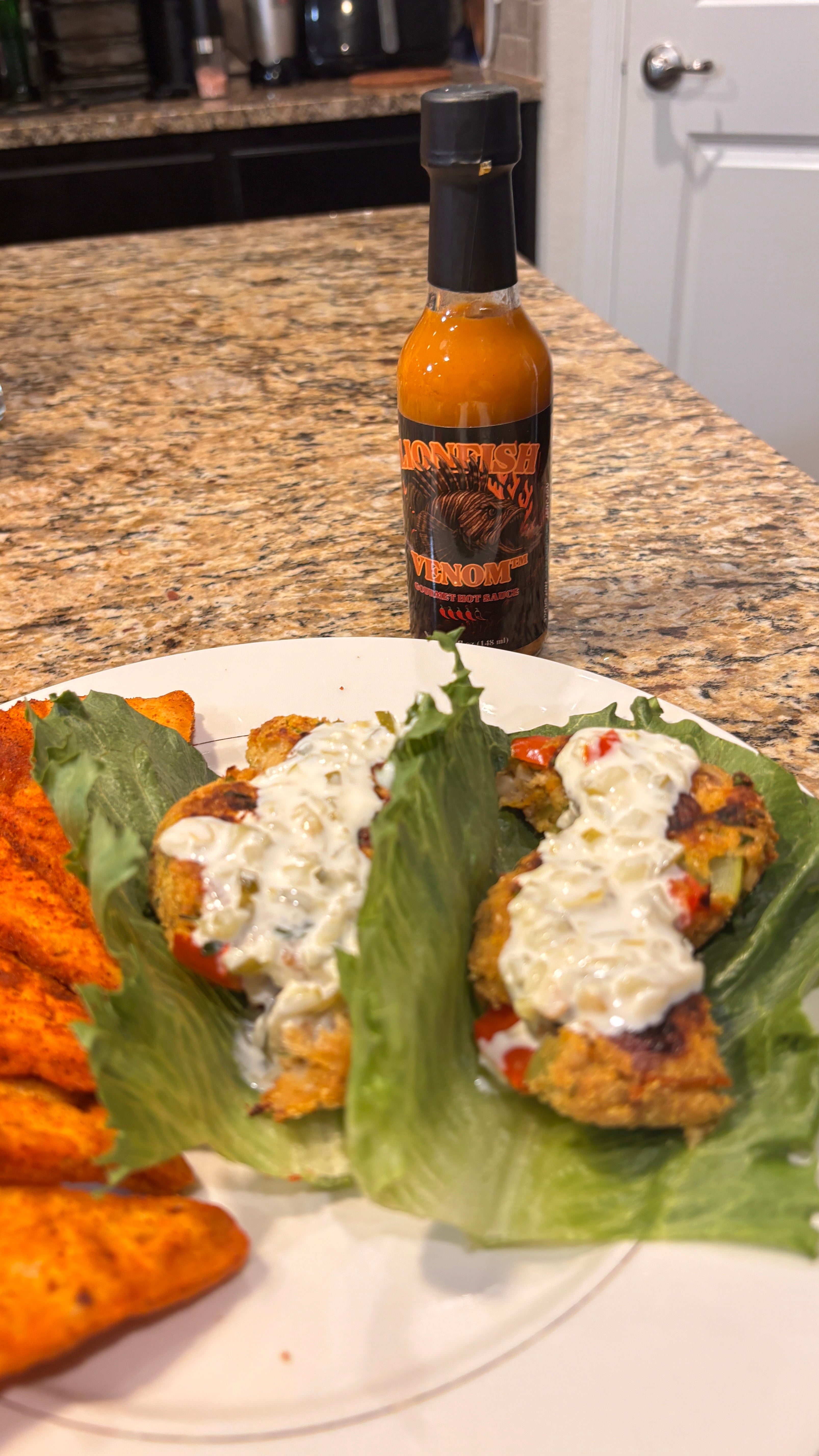 A bottle of Warheads Pepper Sauce stands on a granite kitchen countertop behind a plate of food, including lettuce wraps topped with white sauce and orange sweet potato fries.