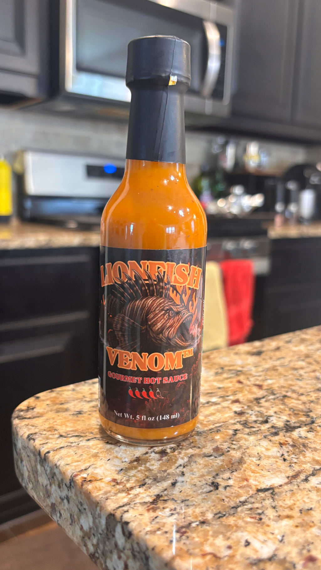 A bottle of Lionfish Venom Hot Sauce sits on a granite kitchen countertop with dark cabinets and appliances blurred in the background.